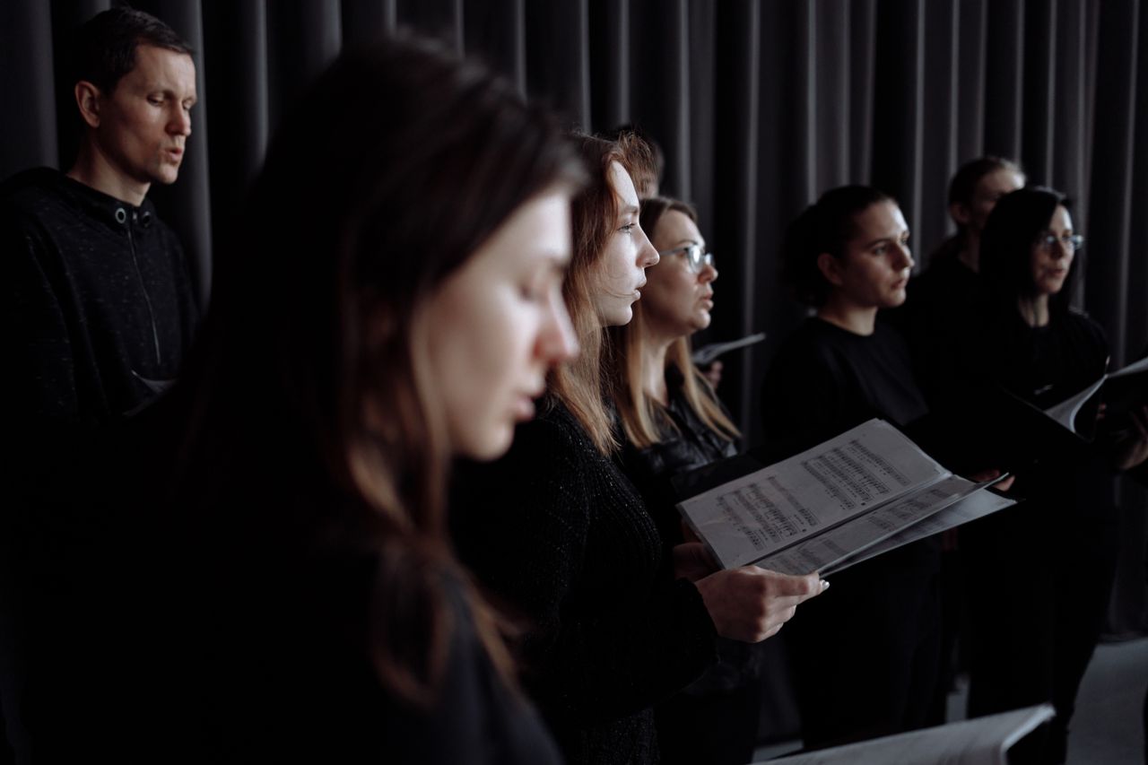 Adult choir practicing in an indoor studio performance setting with music sheets.